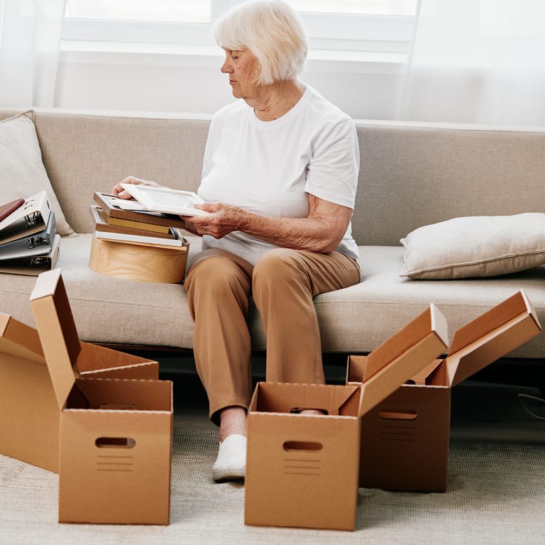 elderly woman sits on a sofa at home with boxes. collecting things with memories albums with photos and photo frames moving to a new place cleaning things and a happy smile. Lifestyle retirement